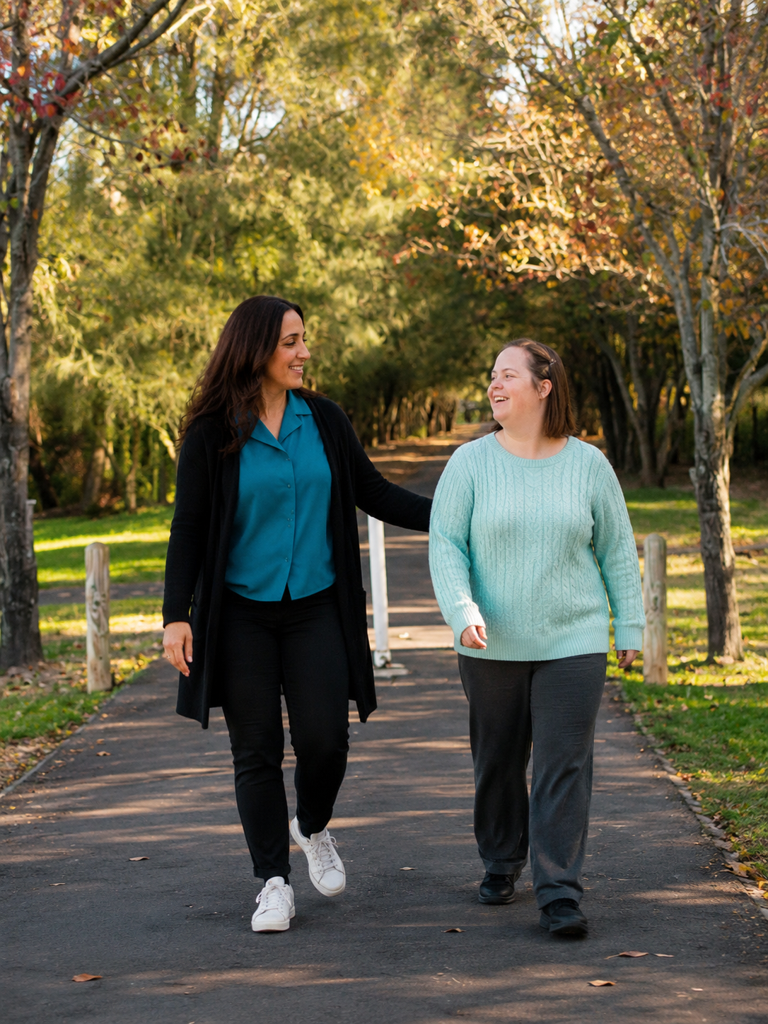 A NutureNest support worker walking with an NDIS participant along a tree-lined park path